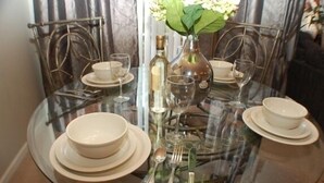 Una mesa de vidrio redonda preparada para cuatro, con platos, fuentes y cubiertos blancos. Un florero con flores está centrado en la mesa.