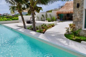 A swimming pool with a white deck, surrounded by palm trees and plants. There is a thatched-roof structure and a stone wall.