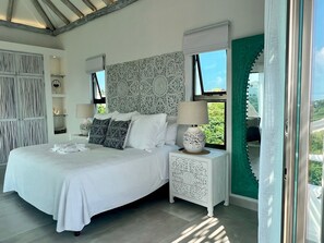 A bedroom with a large bed, white bedding, decorative pillows, a nightstand with a lamp, and a view of greenery outside.