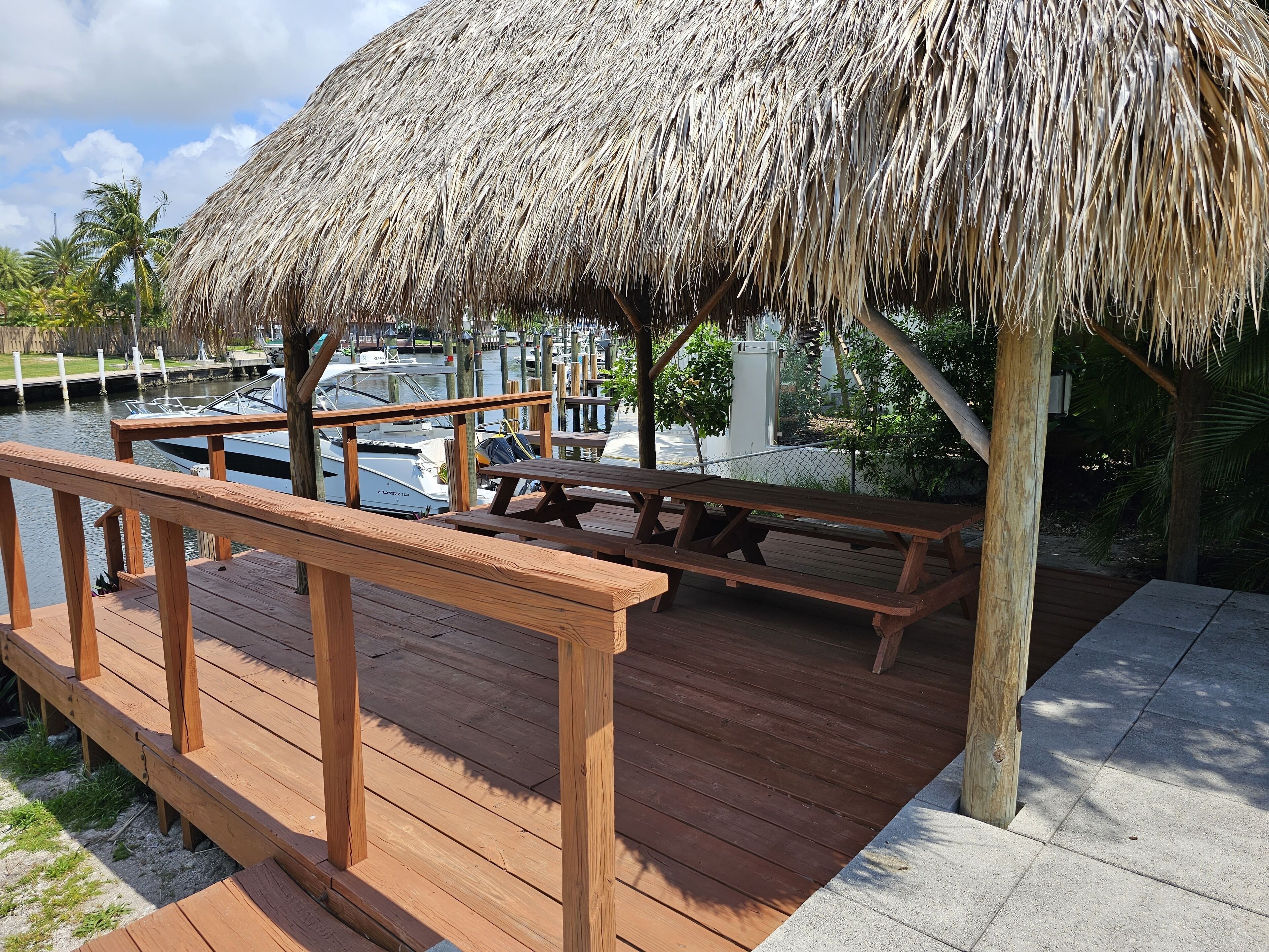 Une terrasse en bois dotée d’une table de pique-nique et de bancs recouverts d’un toit de chaume, offrant une vue sur une marina remplie de bateaux.
