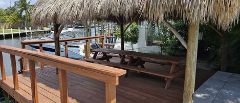 Une terrasse en bois dotée d’une table de pique-nique et de bancs recouverts d’un toit de chaume, offrant une vue sur une marina remplie de bateaux.