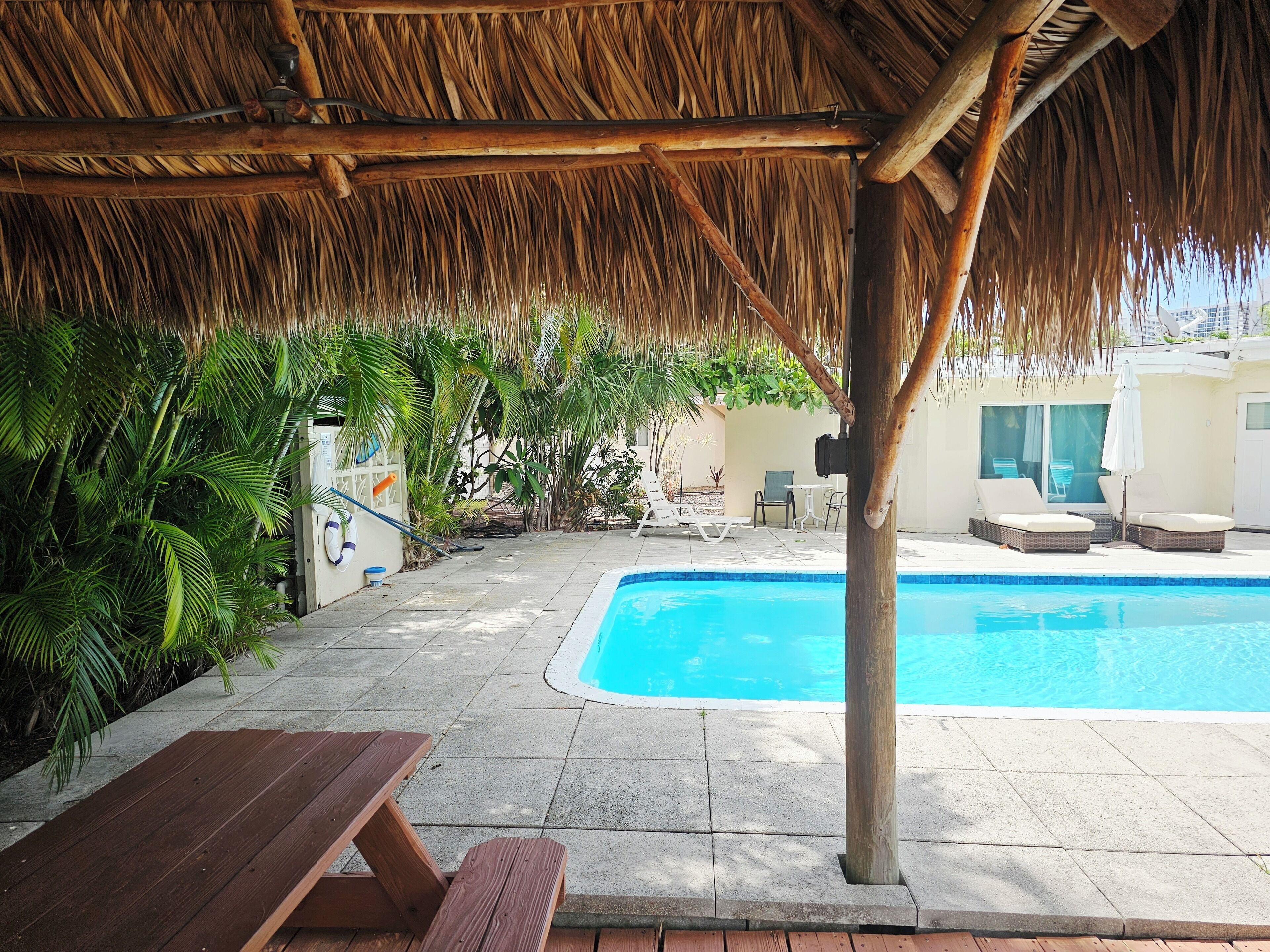 A pool area with a thatched roof, lounge chairs, and a wooden table.