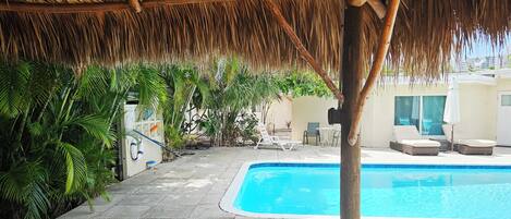A pool area with a thatched roof, lounge chairs, and a wooden table.