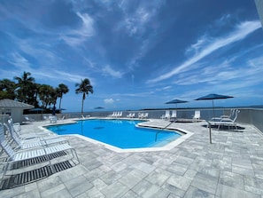 Zona de piscina con sillas de descanso y sombrillas, palmeras y cielo despejado.