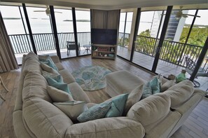 A spacious living room with a large sofa, a flat-screen TV, and a view of the water through large windows.