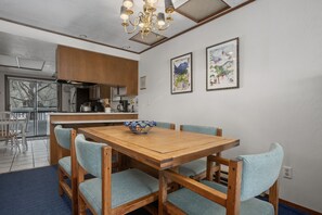 Un coin repas avec une table et des chaises en bois, un lustre, une cuisine en arrière-plan et deux tableaux encadrés au mur.
