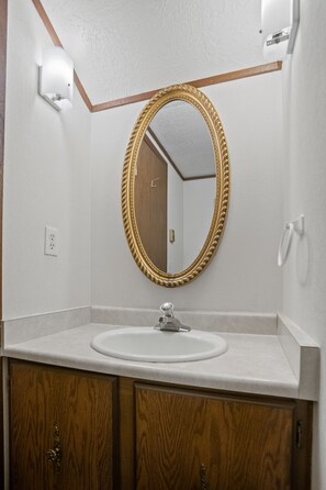 Une salle de bain avec un lavabo blanc, des meubles en bois, un miroir encadré d’or et un mur blanc.