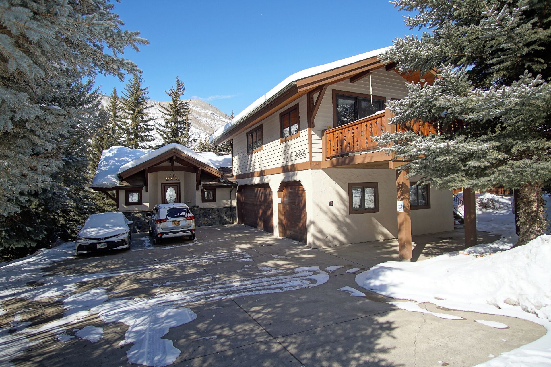 A two-story chalet with a wooden balcony, a garage, and a driveway with snow.