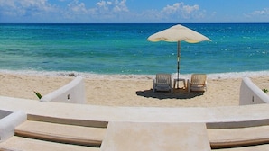 Een strand met een witte parasol en twee stoelen naar de oceaan gericht.