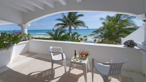 Een overdekt balkon met witte stoelen, een tafeltje met een fles en bloemen, en uitzicht op het strand en palmbomen.