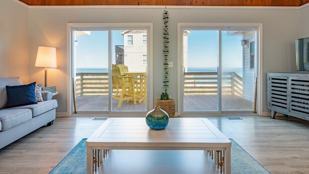 A living room with a wooden ceiling, a white sofa, a table with a vase, and a view of the beach through large sliding doors. - OCS4 Semi-Ocean-Front Home with Gorgeous Views (Nags Head)