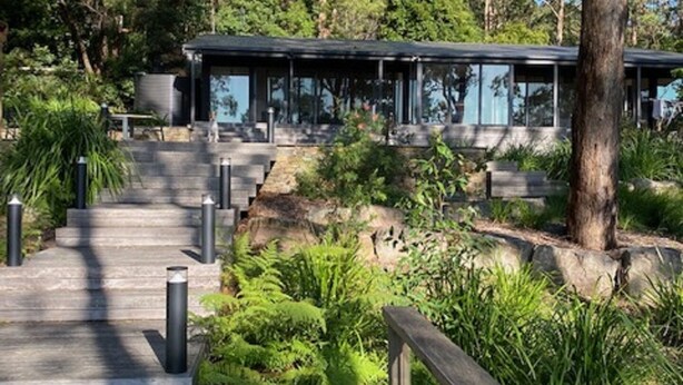 A modern building with a glass facade, surrounded by lush greenery and a wooden pathway leading to it. - Escarpment - Bush to beach views (Berry)