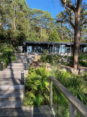 A modern building with a glass facade, surrounded by lush greenery and a wooden pathway leading to it. - Escarpment - Bush to beach views (Berry)