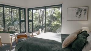 A bedroom with a large bed, a view of trees through large windows, a chair, and a bedside table. - Escarpment - Bush to beach views (Berry)