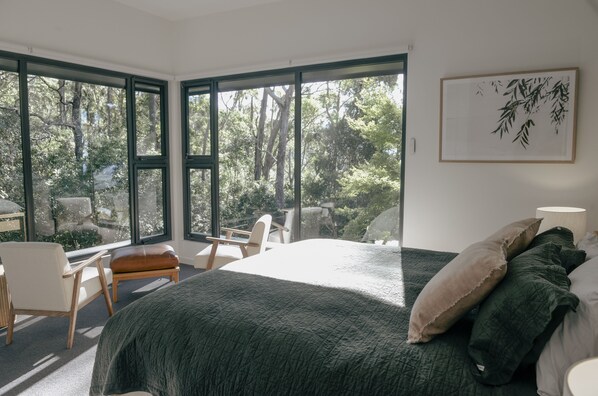 A bedroom with a large bed, a view of trees through large windows, a chair, and a bedside table. - Escarpment - Bush to beach views (Berry)