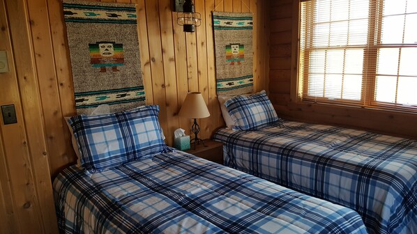 Una habitación con dos camas, paredes de madera, una ventana con persianas y una lámpara en una mesita de noche.