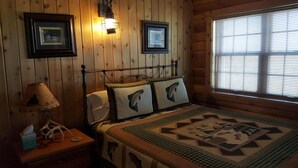 A wooden cabin bedroom with a bed, two pillows with fish designs, a nightstand with a lamp, and two framed pictures on the wall. - Spectacular views and easy access to Fly Fishing on the Madison River (Cameron)