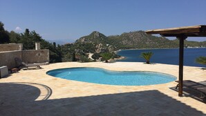 Ein Pool mit geschwungener Kante, umgeben von Steinfliesen. Liegestühle und ein Pergola-Struktur sind vorhanden. Im Hintergrund sind eine Berglandschaft und ein Gewässer zu sehen.