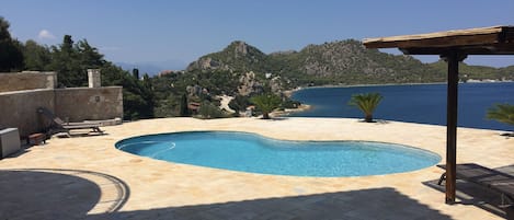 A swimming pool with a curved edge, surrounded by stone tiles. There are lounge chairs and a pergola structure. The background features a mountainous landscape and a body of water.