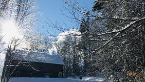 Ein verschneiter Weg führt zu einem Gebäude mit dunklem Dach, umgeben von Bäumen und blauem Himmel.