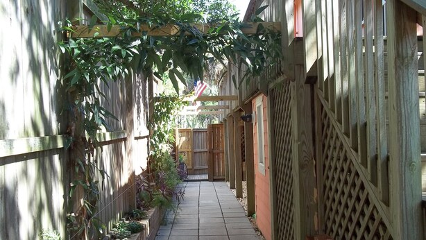 A narrow, paved pathway flanked by wooden fences and vegetation, leading to a wooden structure with a lattice design.