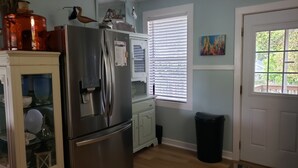 A kitchen with a stainless steel refrigerator, a window with blinds, a trash can, and a door with a view of greenery outside.