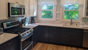 A kitchen with white cabinets, a stove, a microwave, and a coffee maker. There are two windows with white curtains and a view of greenery outside. - 3 BR/2 BA House just steps from Lake Geneva (Williams Bay)