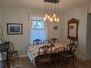 Un coin repas avec une table dressée pour six personnes, entourée de chaises en bois dotées d’assises rembourrées. Un lustre est suspendu au-dessus, et des tableaux encadrés ornent les murs.