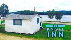 Une petite cabane blanche avec une porte et une fenêtre bleues, portant l’inscription « The Knoll Inn », située au bord d’un lac, devant lequel s’étend une zone herbeuse.