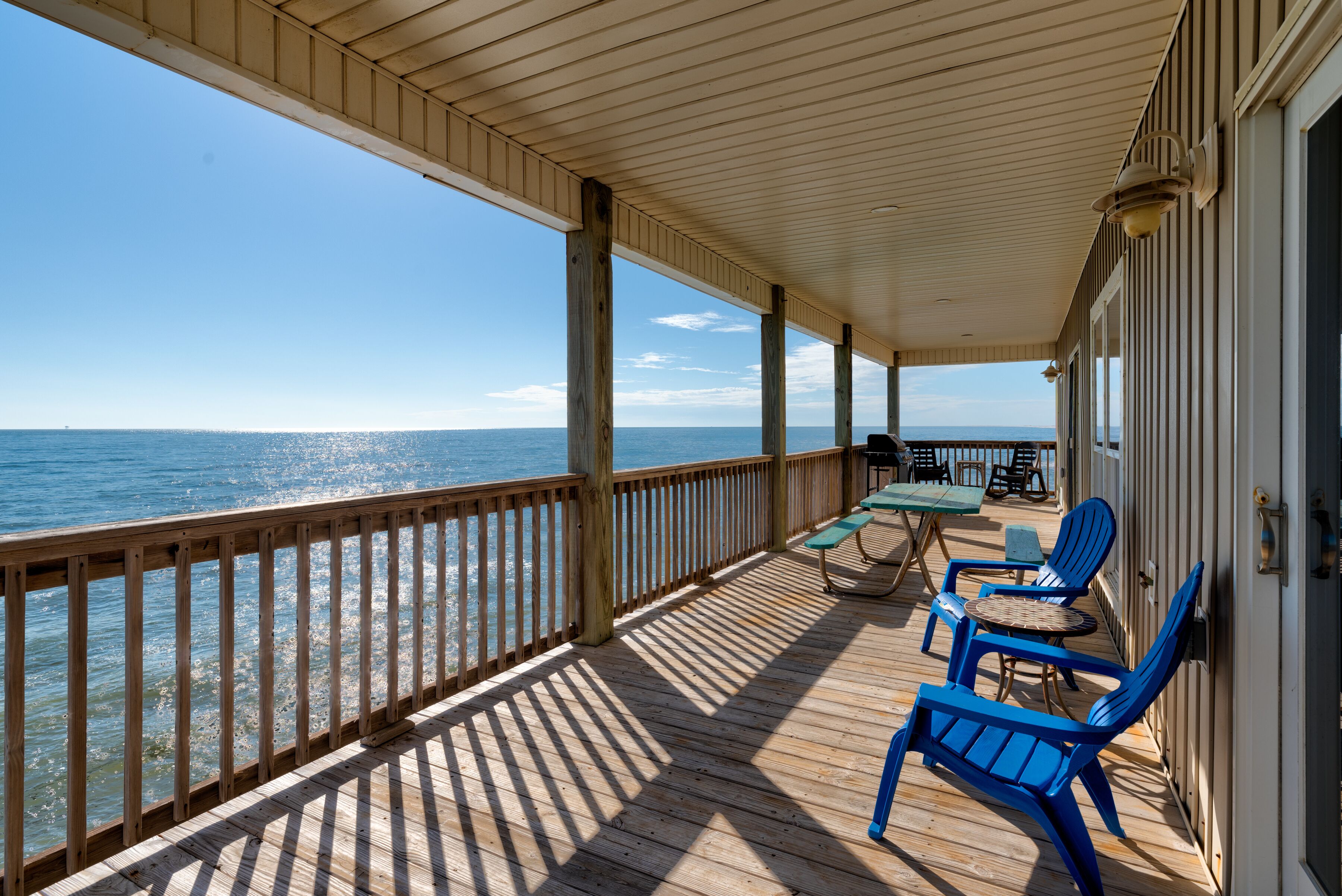A covered deck with wooden railings overlooking the ocean, featuring outdoor seating and a table.