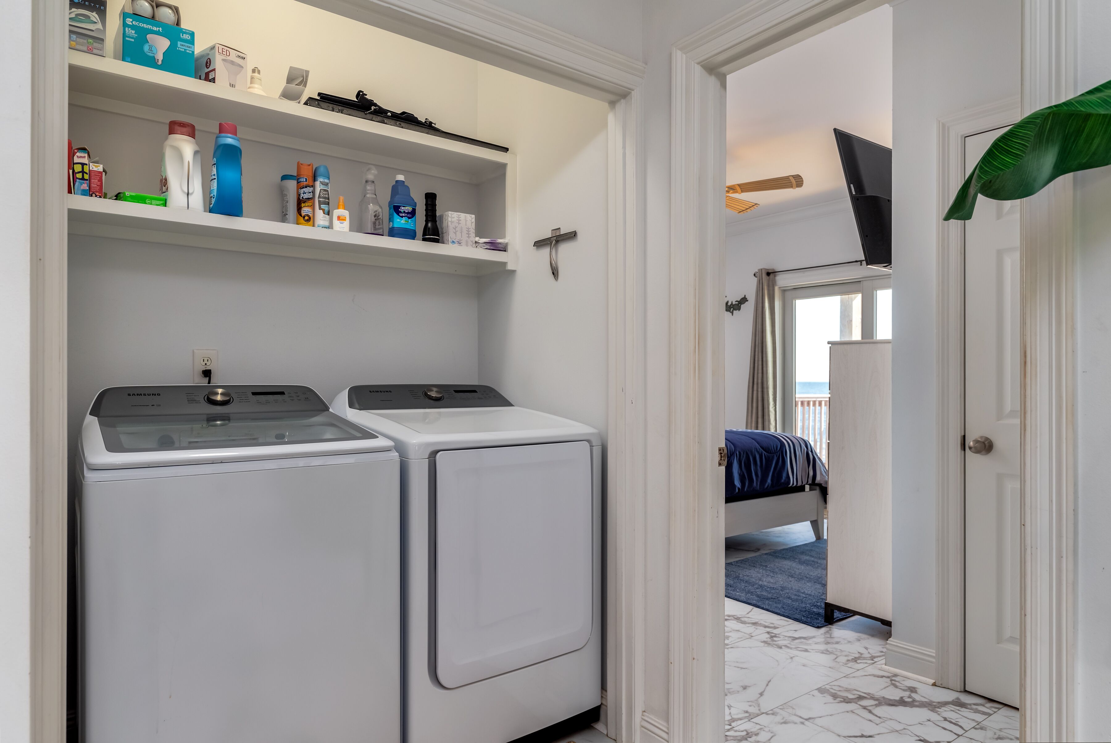 Un'area lavanderia con due lavatrici e asciugatrici bianche, uno scaffale con diversi prodotti per la pulizia e, nella stanza adiacente, un letto con coperta blu.