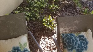 Twee rieten stoelen met bloemenkussens, een kleine tuin met diverse planten en een witte muur met een raam.