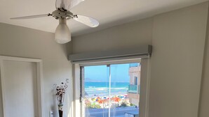 A bedroom with a bed, a ceiling fan, a window with a view of the beach, a small stool, and a shelf with a vase. - Apartment 30 meters from Praia do Forte beach - With beach view. (Cabo Frio)