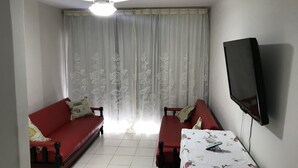 A living room with a red sofa, a small table with a floral tablecloth, a ceiling fan, and a TV mounted on the wall. - Apartment 30 meters from Praia do Forte beach - With beach view. (Cabo Frio)