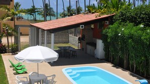 Area piscina con pavimento piastrellato, zona barbecue e posti a sedere all'aperto con ombrellone bianco.