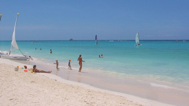 Una spiaggia con acque cristalline turchesi, persone che nuotano e passeggiano, e barche a vela.