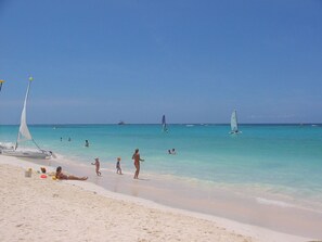 Ein Strand mit klarem türkisblauem Wasser, Menschen beim Schwimmen und Spazierengehen sowie Segelboote.
