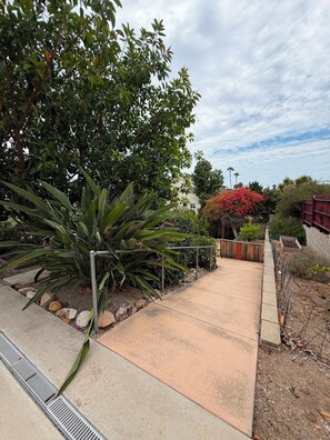 Un chemin pavé mène à un bâtiment, qui est entouré d’une végétation luxuriante et d’un arbuste à fleurs rouges.