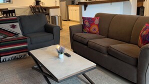 Een moderne woonkamer met een grijze bank, een bruine bank en een salontafel. Achtergrond is een keuken met witte kasten en een klok aan de muur.