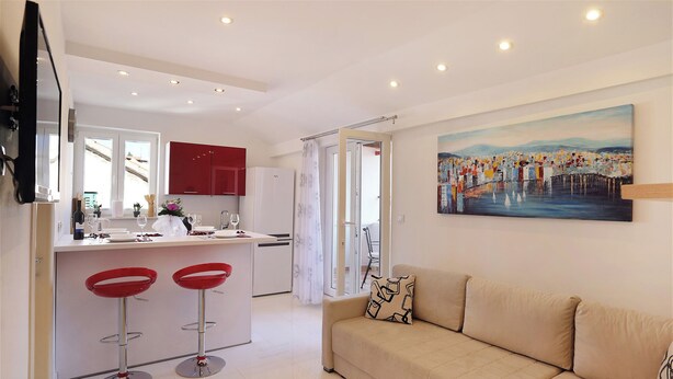 Una sala de estar moderna con un sofá color beige, una zona de comedor con taburetes rojos y una cocina con armarios rojos.