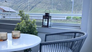 En balkon med et rundt bord, to fletstole, en potteplante og en sort lygt. Udsigt til et bjerg er synlig gennem vinduet.