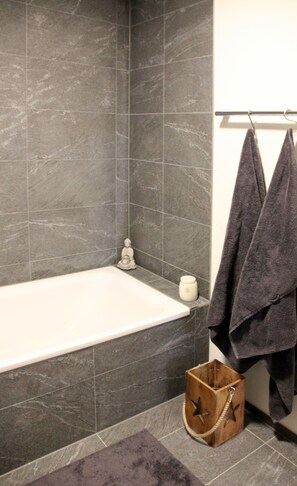 Un bagno moderno con una vasca, pareti piastrellate scure, una piccola statua di Buddha, un portasapone di legno e un portasciugamani con due asciugamani.