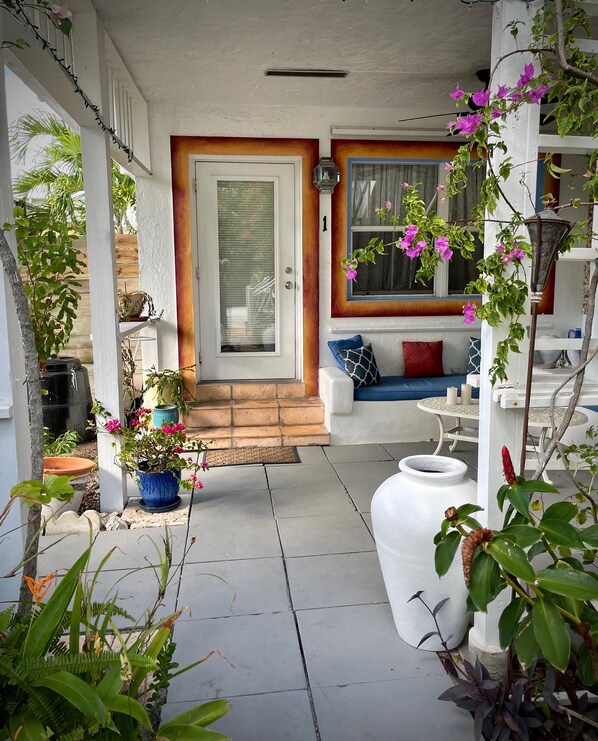 A covered porch with a door, a blue sofa with patterned cushions, and various potted plants. - Located steps from the ocean, offering true beach living. (Hollywood)
