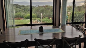 Uma área de refeições com mesa e cadeiras, janelas amplas com vista para vegetação e paisagem ao longe.