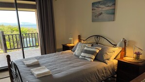 Quarto com cama, duas mesinhas de cabeceira, um criado-mudo, vista para o exterior e um quadro na parede.