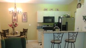 A kitchen with white cabinets, a microwave, and a refrigerator. There is a dining area with a table and chairs. - Spacious one bedroom-one bath condo (Naples)
