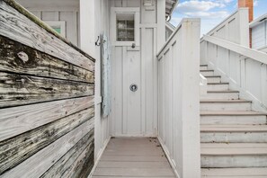 A narrow wooden pathway leading to a white door with a small window, flanked by a white fence and a staircase.
