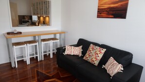 A modern living room with a black leather sofa, white bar stools, a wooden coffee table, and a large window with a view of the sunset. - Ducks Bungalow- Lake Macquarie (Marks Point)