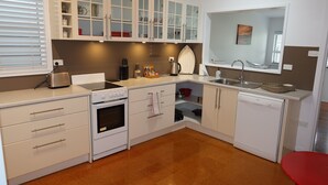 A kitchen with white cabinets, a stove, an oven, a dishwasher, and a red chair. - Ducks Bungalow- Lake Macquarie (Marks Point)