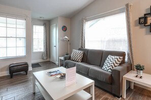 A modern living room with a grey sofa, a white coffee table, and a small side table. There is a window with blinds and a door in the background.